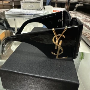 Svarta Saint Laurent solglasögon - Snygga svarta solglasögon från Saint Laurent med breda skalmar och ikoniska YSL-loggor i guld på sidorna. Glasögonen har en fyrkantig form och näskuddar i guld med YSL-detalj. Kommer med originalfodral och putsduk.