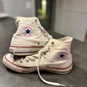 Vita Converse Chuck Taylor All Star - Klassiska vita Converse Chuck Taylor All Star high-tops med röd och blå rand längs sulan. Skorna har snörning, rund tå och är tillverkade i textil med gummisula. Perfekta för dig som gillar en tidlös och cool streetstil. Jag kan stå för frakten. 