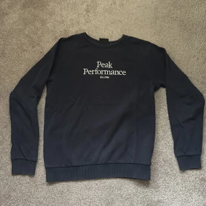 Mörkblå Peak Performance sweatshirt - Snygg mörkblå sweatshirt från Peak Performance med vit broderad logga på bröstet. Tröjan har rund hals, långa ärmar och ribbade muddar vid ärmslut och nederkant. Perfekt för dig som gillar stilren och sportig look. Stl 160