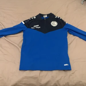 Blå JAKO fotbollströja Eskilstuna City - Sportig blå och svart långärmad tröja från JAKO med Eskilstuna City-tryck på ryggen och klubbmärke fram. Halv dragkedja framtill och vita detaljer på axlarna. Perfekt för fotbollsträning eller som supportertröja.