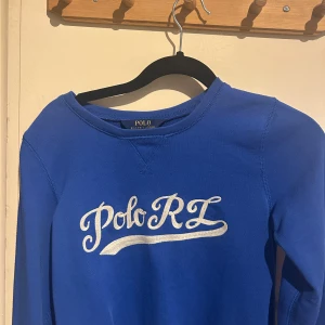 Blå tröja från Polo Ralph Lauren - Snygg blå tröja från Polo Ralph Lauren med vit broderad text 'Polo RL' på bröstet. Klassisk rund halsringning och långa ärmar. Perfekt för dig som gillar stilrena och sportiga plagg med ikonisk logga. Hör av dig om du undrar något😁