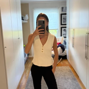 Vit ribbad ärmlös topp med knappar - Säljer en vit ribbad topp med v-ringning och tre dekorativa guldknappar framtill. 