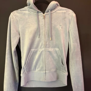 Ljusblå velour hoodie Juicy Couture - Mjuk och lyxig ljusblå hoodie från Juicy Couture i velour med dragkedja, huva och snörning. Tröjan har känguruficka och ribbade muddar. Perfekt för en chill och trendig look. en metallsak lossnat