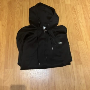 Svart hoodie från Lacoste  - Svart hoodie från Lacoste med klassisk krokodillogga på bröstet. Tröjan har huva med snörning, hel dragkedja framtill och ribbade muddar. Perfekt för en chill och stilren look. Materialet är mjukt och känns skönt mot huden.