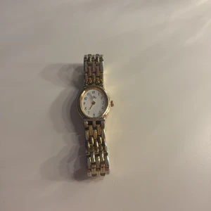 Guld klocka - Snygg klocka med rund vit urtavla och tydliga svarta siffror. Armbandet är i metall med guldfärgade länkar som ger en schysst kontrast. Klassisk design med metallarmband och stilren look, perfekt för dig som gillar tidlösa accessoarer. Klockan fungerar så man kan kolla tiden på den.