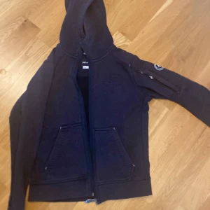 Marinblå hoodie från Pelle P med huva - Marinblå hoodie från Pelle P med dragkedja, broderad logga på bröstet och patch på ärmen. Tröjan har huva, långa ärmar och två fickor framtill. Perfekt för dig som gillar en sportig och clean stil.