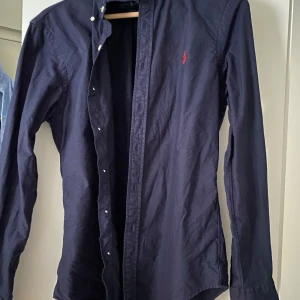 Mörkblå skjorta från Ralph Lauren - Snygg mörkblå skjorta från Ralph Lauren i slim fit-modell. Skjortan har långa ärmar, klassisk krage och knappar framtill. På bröstet finns det ikoniska röda Polo-broderiet. Materialet är mjuk bomull som känns skönt mot huden.