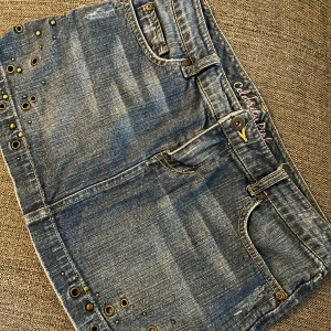 Jeanskjol med nitar från Celebrity Pink - Snygg kort jeanskjol från Celebrity Pink i blå denim med slitningar och coola guldfärgade nitar längs kanterna. Kjolen har klassiska fickor fram och bak, bälteshällor och broderad logga i rosa vid linningen. Perfekt för dig som gillar en edgy stil.