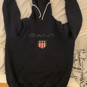 Marinblå hoodie från GANT - Säljer en marinblå hoodie från GANT med vit snörning i huvan och klassiskt broderat GANT-logo samt sköld med stjärnor och ränder på bröstet. Stor känguruficka framtill och mjukt material, perfekt för chill dagar.