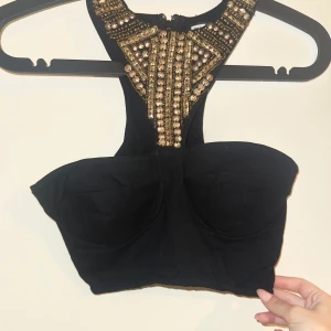 Svart korsettopp med guldpärlor - Supersnygg svart korsettopp med breda axelband och detaljer i guldpärlor och paljetter framtill. Toppen har markerade kupor och dragkedja i ryggen för en tight passform. Perfekt för dig som vill sticka ut med en edgy och glam look.