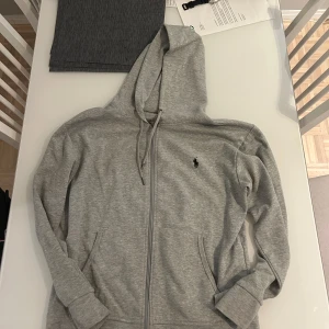 Grå hoodie från Polo Ralph Lauren - Grå hoodie från Polo Ralph Lauren med dragkedja framtill, huva med snörning och klassisk logga broderad på bröstet. Tillverkad i mjuk bomullsmix, perfekt för chill dagar. Långa ärmar och två fickor framtill. I bra skick och den ät Äkta!