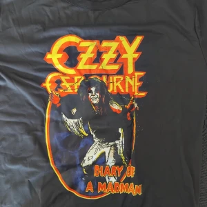 Ozzy Osbourne Diary of a Madman t-shirt - Svart t-shirt med färgstarkt tryck av Ozzy Osbourne och texten 'Diary of a Madman' i röd och gul. Motiv från albumet, med cool retrostil och stor grafik på framsidan. Perfekt för dig som älskar rock och vill sticka ut med en unik bandtröja.