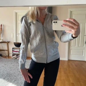 Grå hoodie från Adidas med dragkedja - Säljer en grå hoodie från Adidas med klassiska vita ränder längs ärmarna och hel dragkedja framtill. Tröjan har huva, ribbade muddar och är tillverkad i mjuk bomullsblandning. Perfekt för en sportig och avslappnad stil.