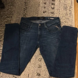 Replay Anbass jeans mörkblå, stl 29 - Snygga mörkblå Replay Anbass jeans med klassisk femficksdesign och diskreta slitningar. Jeansen har raka ben, normal passform och är tillverkade i mjukt denimtyg med stretch för extra komfort. Perfekta för dig som gillar stilrena och bekväma jeans.