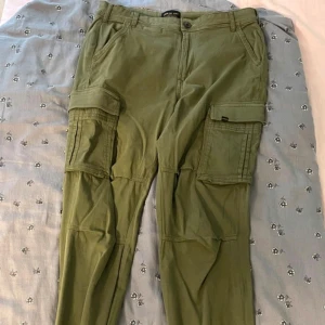 Olivgröna cargopants från Denim Day - Snygga olivgröna cargopants från Denim Day med klassiska stora fickor på sidorna och raka ben. Byxorna är tillverkade i bomull och har en avslappnad passform med bälteshällor och dragkedjegylf. Perfekta för en chill och streetig look.