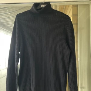 Svart ribbad polotröja - Svart ribbstickad polotröja med lång ärm och hög krage. Tröjan är i ett mjukt, stickat material och har en klassisk, clean look som funkar till allt. 