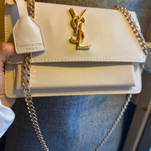 Vit axelväska från Saint Laurent - Snygg vit axelväska från Saint Laurent med guldfärgad kedja och ikoniskt YSL-emblem framtill. Väskan är i skinn och har en stilren, rektangulär form med lock och detaljerad sömnad. Perfekt för dig som vill ha en lyxig och trendig accessoar.helt ny