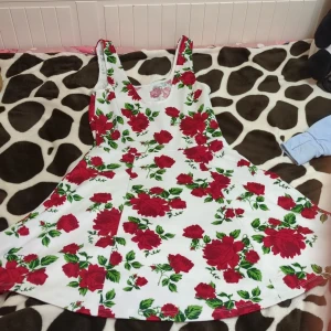 Vit blommig klänning från Divided - Vit klänning från Divided med röda rosor och gröna blad över hela tyget. Klänningen är ärmlös, har rundad halsringning och en utställd kjol som ger en söt och somrig vibe. Priset kan diskuteras.