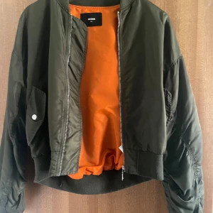 Grön bomberjacka med brodyr Bik Bok - Snygg mörkgrön bomberjacka från Bik Bok med orange foder och broderad text 'WASTE NO TIME' i svart och orange på ryggen. Jackan har dragkedja framtill, fickor med tryckknapp och är långärmad med ribbade muddar.