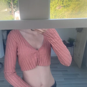 Rosa croppad ribbad topp från Monki - Säljer en croppad långärmad topp från Monki i en ljusrosa nyans. Toppen är ribbstickad med v-ringning och knappar framtill. I storlek s.