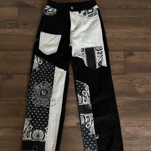 Patchwork jeans i svart och vitt - Unika patchwork jeans i svart och vitt med bandana-mönster och kontrasterande tygbitar. Raka ben, flera fickor och coola detaljer som ger en edgy streetkänsla. Perfekta för dig som vill sticka ut med din stil.