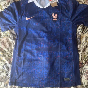 Blå Frankrike fotbollströja Nike - Säljer en officiell Frankrike fotbollströja från Nike med korta ärmar. Tröjan har ett digitalt mönster i olika blå nyanser med små röda detaljer och vit krage. FFF-logga med tupp på bröstet och Nike swoosh i guld. Materialet är lätt och andas, perfekt för match eller träning.