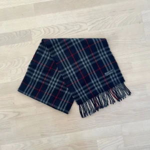 Rutig halsduk från Burberrys i lammull - Snygg rutig halsduk från Burberrys of London i 100% lammull. Mörkblå bas med grå och röda rutor, klassisk design och fransar i ändarna. Perfekt accessoar för att lyfta din outfit med en tidlös brittisk touch.