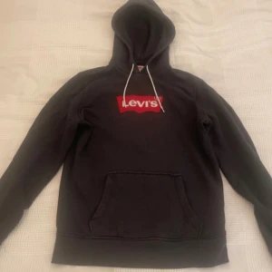Svart Levi's hoodie med röd logga - Svart hoodie från Levi's med klassisk röd logga på bröstet. Tröjan har huva med vita snören och en stor magficka. Tillverkad i mjuk bomullsblandning med 10% polyester. Perfekt för en chill och stilren look.