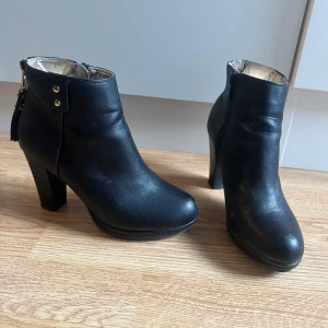 Svarta stövletter med klack och dragkedja - Säljer ett par svarta stövletter i skinnimitation med hög klack och dragkedja bak och sida. Snygga detaljer med nitar och rund tå. Perfekta för dig som vill ha en stilren och edgy look. Klacken är 9,5cm