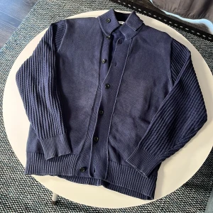 Mörkblå kofta från Nano Universe - Säljer en mörkblå kofta från Nano Universe i storlek L. Koftan har ribbade ärmar och muddar, hög krage och knäppning med svarta knappar framtill. Materialet är en mix av bomull och polyester, vilket gör den både mjuk och slitstark.