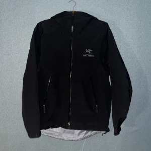 Svart vindjacka från Arc'teryx M - Snygg svart vindjacka från Arc'teryx i storlek M. Jackan har huva, hel dragkedja framtill och två dragkedjeförsedda fickor. Materialet är lätt och känns slitstarkt, perfekt för blåsiga dagar. Broderad logga på bröstet ger en clean och sportig look. Priset kan diskuteras vid snabb affär.