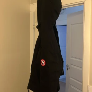 Svart dunjacka från Canada Goose - Säljer en svart dunjacka från Canada Goose med klassisk patch på ärmen och huva. Jackan har dragkedja framtill och är fodrad för att hålla dig varm under kalla dagar. Perfekt för dig som vill ha en stilren och ikonisk vinterjacka.kommer inte till användning längre, därför säljer jag den.