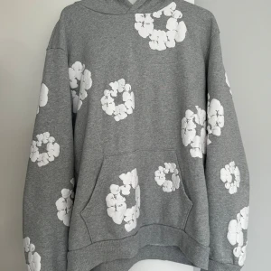 Grå hoodie med vita blommor Denim Tears - Grå Denim Tears Cotton Wreath Hoodie i mjuk bomull, oversized passform med huva och känguruficka. Ikoniskt bomullskrans-tryck runt hela hoodien – en av märkets mest eftertraktade modeller. Snygg, bekväm och svår att få tag på här i Sverige!  Hoodien har inga skavanker. Den är använd ett x antal gånger men är i bra skick. Materialet är inte typiskt mjukt som man tror utan lite ”hårdare”. Men den är i bra skick.   Nypris ca 5000kr, mitt pris är 1500kr