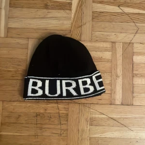 Svart mössa från Burberry - Säljer en svart mössa från Burberry med vit logotyp runt hela kanten. Klassisk och stilren design, perfekt för dig som vill ha något snyggt och enkelt. Mössan är stickad och har en mjuk känsla.