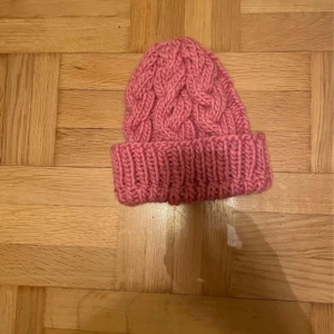 Rosa stickad mössa med flätmönster - Supermysig rosa mössa med bred uppvikt kant och snyggt flätstickat mönster. Perfekt för kalla dagar och ger en cool vibe till din vinterstil. Tjockt garn som håller dig varm och ger en trendig look.