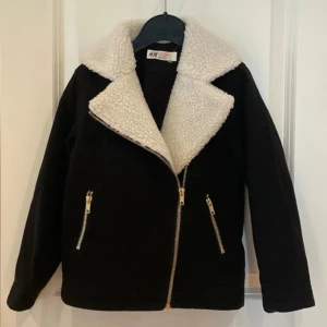 Svart teddyjacka från H&M med dragkedja för barn - Svart teddyjacka från H&M med stor vit fuskpäls-krage och två snedställda fickor med guldiga dragkedjor. Jackan har en schysst bikerstil och är perfekt för kyliga dagar. Materialet är mjukt och mysigt, och modellen är normal i passformen.