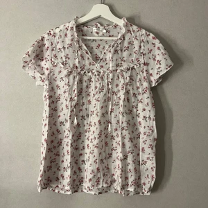 Blommig vit blus från Angelology - Supersöt blus i vitt från Angelology med små röda blommor och gröna blad. Blusen har korta ärmar, v-ringning med knytband och volangdetaljer vid halsen. Tunt, luftigt tyg i bomull som passar perfekt till varmare dagar. Pris kan diskuteras🤗