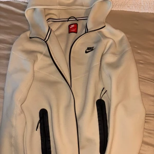 Vit Nike tech kofta  - Säljer en stilren vit nike tech från Nike med svarta dragkedjor och svarta detaljer längs kanterna. Tröjan har huva och två fickor med dragkedja framtill. Perfekt för en sportig och avslappnad look.