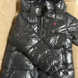 Svart pufferjacka från Polo Ralph Lauren - Säljer en svart glansig pufferjacka från Polo Ralph Lauren med röd broderad logga på bröstet. Jackan har dragkedjor framtill och på fickorna, quiltad design och huva. Perfekt för kalla dagar och riktigt snygg streetstil. Jackan är använd ett par gånger och är som ny. Storleken S men jag har storlek M i tröjor och det passar perfekt