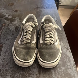 Gråa Vans Old Skool sneakers - Säljer ett par klassiska Vans Old Skool sneakers i grått med vit sula och vit sidostrip. Skorna har snörning, rund tå och platt gummisula med det ikoniska rutmönstret undertill. Ovandelen är i canvas och mocka, insida i vitt syntetmaterial. Perfekta för en avslappnad streetstil. Storlek 40 