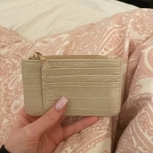 Beige plånbok med krokoprägling - Snygg beige plånbok i croco-imitation med flera kortfack och guldfärgad dragkedja. Slimmad och stilren design som passar perfekt i fickan eller väskan. Perfekt för dig som gillar minimalistisk och trendig stil. Lite av de guldiga vid öppningen har försvunnit man kan se det på bild 2. Plånboken har använts endast några enstaka gånger. (Denna säljs igen då personen inte hämtade ut sitt paket och fick tillbaka den i retur)