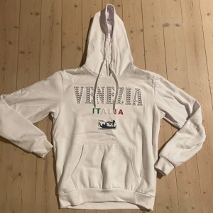 Hoodie vit  - Pris kan diskuteras, skickar gärna bilder på tröjan om ni vill ha! Vit hoodie med tryck 'Venezia Italia' i grått och färgglada bokstäver samt gondolmotiv. Tröjan har huva med snörning, magficka och långa ärmar. Perfekt för dig som gillar streetstyle och vill ha en chill look.