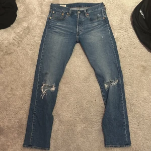 Levi's 501 blå jeans med slitningar - Klassiska blå Levi's 501 jeans med raka ben och snygga slitningar på båda knäna. Jeansen har fem fickor, knappgylf och den ikoniska läderpatchen bak i midjan. Perfekta för en avslappnad och trendig look.