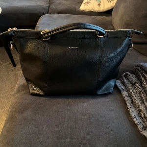 Svart handväska från Puccini i skinn - Helt oanvänd svart handväska från Puccini i skinn med guldfärgade detaljer. Väskan har dragkedja upptill, ett ytterfack med dragkedja och ett rymligt, mönstrat innerfoder med flera innerfoder. Funkar jätte bra till skola och jobb då den får plats med större datorer och böcker. 