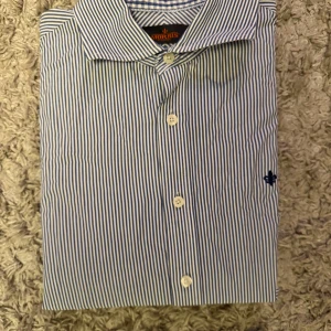 Randig skjorta från Morris - Snygg skjorta från Morris med smala blå och vita ränder, klassisk krage och knäppning framtill. Broderad logga på bröstet. Skjortan har lång ärm och normal passform, perfekt för en clean och stilren look. Slim fit