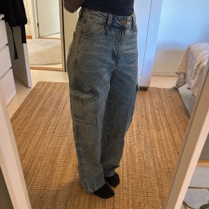 Blå cargojeans med bred passform - Säljer ett par blå cargojeans med hög midja och bred passform. Jeansen har stora sidofickor och klassiska bakfickor. Tillverkade i denim med en avslappnad, loose fit och raka ben. Perfekta för en chill och trendig look.