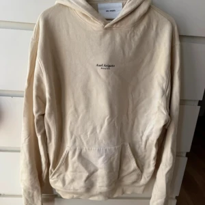 Beige hoodie från Axel Arigato - Snygg beige hoodie från Axel Arigato med minimalistisk logga på bröstet. Tröjan har huva, känguruficka och är tillverkad i 100% bomull för en skön och avslappnad känsla. Perfekt för dig som gillar stilrena och trendiga plagg.