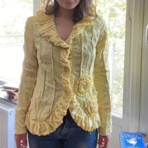 Gul volangkofta från L33 - Superfin gul kofta från L33 med volangdetaljer längs krage, knappslå och nederkant. Koftan har långa ärmar, dekorativa blommor framtill och ett skrynklat tyg som ger en cool look. Perfekt för dig som vill sticka ut med färg och detaljer.