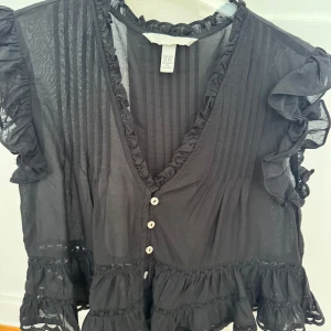 Svart volangblus från H&M - Supersöt svart blus från H&M med volanger på ärmar och nederkant, v-ringning med knappar framtill och tunna ränder i tyget. 