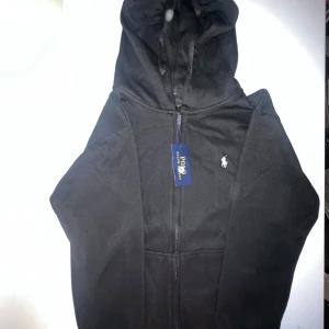 Svart hoodie från Polo Ralph Lauren - Svart hoodie från Polo Ralph Lauren med dragkedja framtill och klassisk huva med snörning. Tröjan har det ikoniska vita Polo-logot broderat på bröstet och är tillverkad i mjuk bomullsmix. Perfekt för en chill och stilren look.